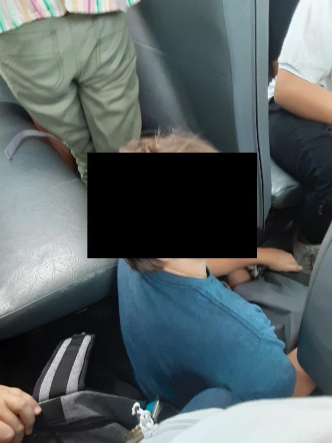 A student sits on the floor of a bus while all of the seats are full. (Student did not want their face shown).